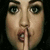 A close-up of a person's face with a finger pressed to lips in a shushing gesture, set against a dark background.