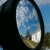 Pixelated oval-shaped icon with black border showing a landscape reflection of blue sky, white clouds, and green-brown terrain.