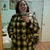 A pixelated figure in a dark green and brown plaid shirt stands against a blurred background with light blue and beige tones.
