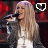 A pixelated image of a woman with long blonde hair singing into a microphone, wearing a white top and dark jacket against a red background.