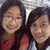 Two people with dark hair pose closely together in a pixelated static image, one wearing a red garment, the other in a light-colored top against a blurred background.