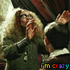 A woman with curly blonde hair and glasses raises her hands in a theatrical pose, wearing a dark green jacket over a white-collared shirt.
