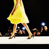A person in a yellow dress and high heels walks on a white surface against a dark background with blurred lights.