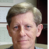 Static pixelated image of a person with light brown hair, visible ear, and partial dark clothing against blurred background elements.