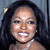 A pixelated close-up of a person with dark hair, light skin, and a silver necklace, smiling with visible teeth against a dark background.