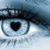 A pixelated close-up of a stylized eye with a black heart-shaped pupil, set against a blurred blue-gray background.