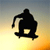 Black silhouette of a skateboarder performing a trick against a gradient sunset background with yellow-to-orange hues at the bottom transitioning to blue-gray at the top.