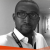 Pixelated grayscale portrait of a person wearing glasses and a white collared shirt with a dark tie, set against a light gray background with an orange horizontal bar at the bottom.