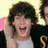 A person with curly brown hair wearing a black jacket over a white shirt, smiling broadly with mouth open and hand near head against pink and yellow background.