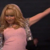 A person with long blonde hair wearing a light pink sleeveless top with small dark dots and a silver sequined shoulder strap, arm extended to the right against a dark background.