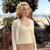 A pixelated figure with light hair wears a white long-sleeve top and light-colored bottoms, standing outdoors with trees and a bright sky in the background.