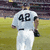 A baseball player in a white uniform with the number 42 on his back walks across a grassy field, viewed from behind.