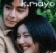 Two people in close proximity with dark hair and light-colored clothing against a green background, text 'k.mayo' visible above.