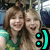 Two people with brown and blonde hair smiling, wearing green clothing, next to a cyan smiley face symbol on a pixelated background.