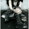 A person in a black sleeveless top and layered dark skirt sits with knees drawn up, hands clasped over one knee, wearing black boots and a choker necklace.