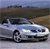 A silver sedan with black wheels and dark-tinted windows is positioned on a light gray surface against a blurred background of brown and blue tones.