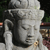 Static pixelated image of a stone or marble statue head with closed eyes, dark hair, and a smooth surface, set against a blurred background with green and brown tones.