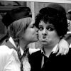 Black-and-white image of two people in close proximity, one kissing the other's cheek while the other looks directly at the viewer with a surprised expression.