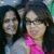 Two people with dark hair pose closely together; one wears a pink garment, the other light blue. The background is blurred and indistinct.