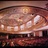 A pixelated static image showing a blurred interior view of a theater with rows of seats and a stage area under warm lighting.