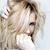 Pixelated image of a person with light hair wearing a white top, holding a small object near their face against a light background.
