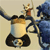 Pixelated static image shows a cartoon character in red shirt and brown pants standing on a vehicle with large black tires and a yellow top, next to a blue figure with raised arm.