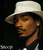 Pixelated static image of a person wearing a light-colored wide-brimmed hat and dark clothing against a black background, with visible dithering and low-resolution rendering artifacts.