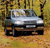 Static pixelated image of a light blue sedan with dark gray accents parked on a brown dirt path, surrounded by blurred green and brown foliage.