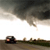 A static image shows a dark vehicle on a road under a cloudy sky with a bright horizon line.