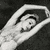 Black-and-white image of a ballet dancer in mid-pose with arms extended and one leg lifted, wearing a short skirt and pointe shoes.