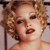 Pixelated close-up of a person with curly light brown hair, dark eyes, and red lips, wearing a dark strap over the shoulder against a light background.