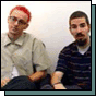 Two men sit side-by-side; the man on the left has red hair and glasses, while the man on the right has dark hair and a beard.