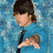A person in a blue shirt with dark hair and bangs, making a hand gesture against a textured blue background.
