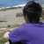 A person in a purple shirt sits with back to camera, facing beach and ocean under blue sky.