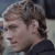 Pixelated close-up of a person's head and shoulder in profile, showing light brown curly hair, a dark top, and a blurred background.