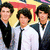 Three young men in formal attire stand together against a yellow and red background with an orange 'R' symbol on black.