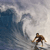 A pixelated image shows a surfer riding a large wave under a cloudy sky, with the surfer wearing a brown top and dark shorts.