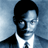 Pixelated portrait of a person with short dark hair wearing a dark suit jacket, white shirt, and dark tie against a blue gradient background.
