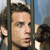 A pixelated close-up of a person's face behind vertical bars, with a blurred background showing partial figures and a blue sky.