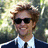 A person with light brown curly hair wearing dark sunglasses, a white collared shirt, and a black tie against a blurred green background.