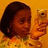 A person with dark hair holds a light-colored phone against an orange background, wearing a patterned shirt.