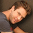 A pixelated image of a person with brown hair wearing a gray shirt, lying down with one arm behind their head and the other resting on their chest.