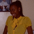 A person with dark skin and short hair wears a yellow shirt with a brown symbol, standing against a light background.
