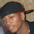 Pixelated close-up of a person wearing a dark cap with red trim, light skin tone, and visible ear, against a blurred background.