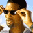 Pixelated image of a person with dark hair wearing sunglasses, holding a light-colored object near their face against a light blue background.