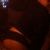 Dark brown figure with orange accents in a dimly lit setting, featuring a small blue pixel cluster in the top-left corner.
