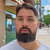 Pixelated static image of a person with dark hair and beard wearing a light-colored shirt against a blurred background with green and blue tones.