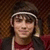 Pixelated static image of a person wearing a dark hat with a light band, dark sunglasses, and a red and dark-colored top against a brown textured background.