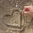 A hand draws a heart in sand with text reading 'i love you' across four