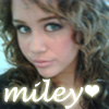 A close-up portrait of a person with curly brown hair and light blue eyes, wearing a dark top with a glowing white text overlay reading 'miley' and a heart symbol.